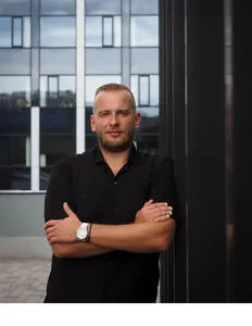 Modrá minimalistická ikona domečku s lupou, skrze lupu je vidět symbol dolaru.