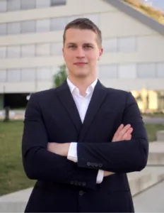 Modrá minimalistická ikona domečku s lupou, skrze lupu je vidět symbol dolaru.